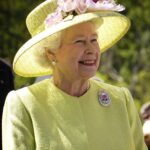 Her Majesty Queen Elizabeth II shown smiling in a yellow green suit and hat adorned with flowers
