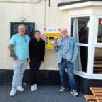 Dennis Frith shown on the right with a well-deserved pint in front of the defibrillator bought with money raised through a campaign with The Rumbold Arms in Great Yarmouth